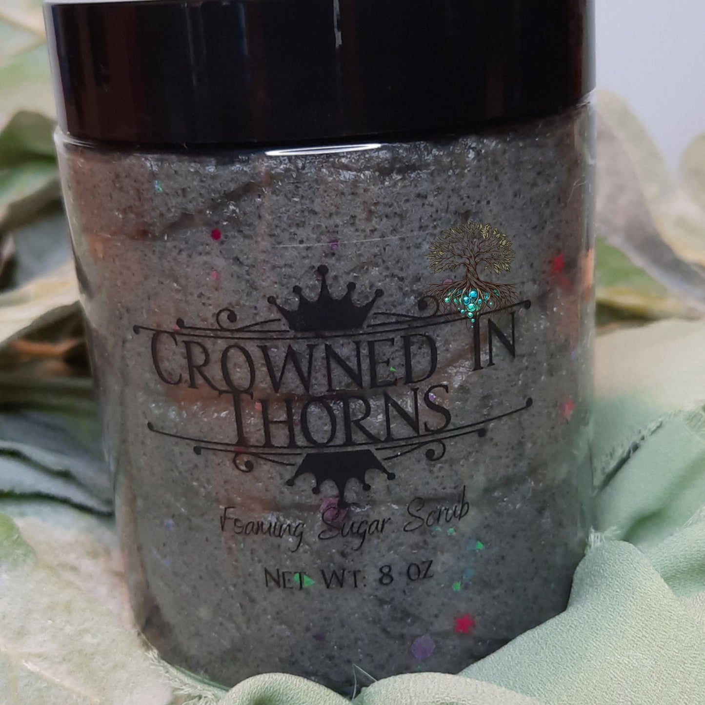 Jar of 'Crowned in Thorns' foaming sugar scrub with decorative elements on a green leafy background.