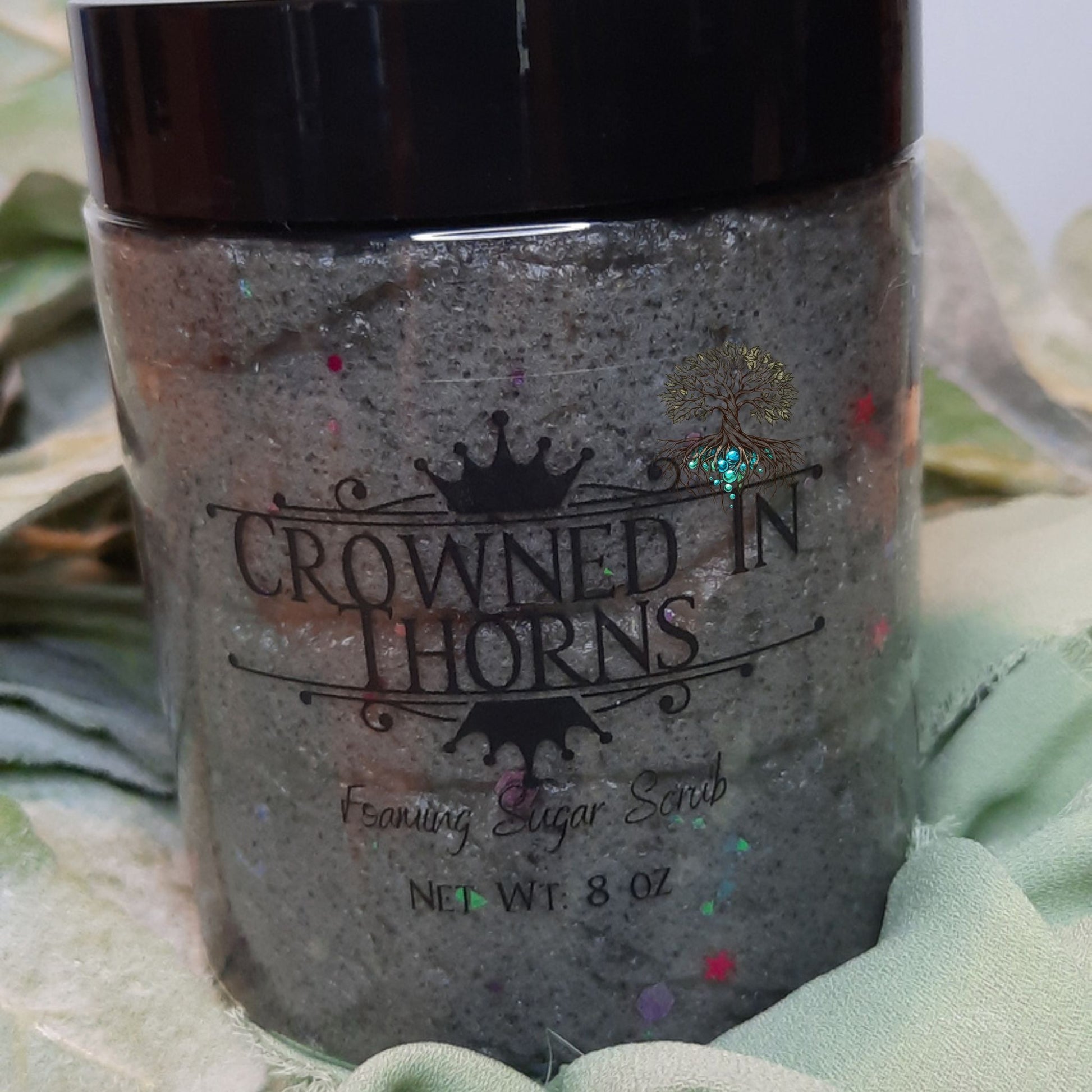 Jar of 'Crowned in Thorns' foaming sugar scrub with decorative elements on a green leafy background.